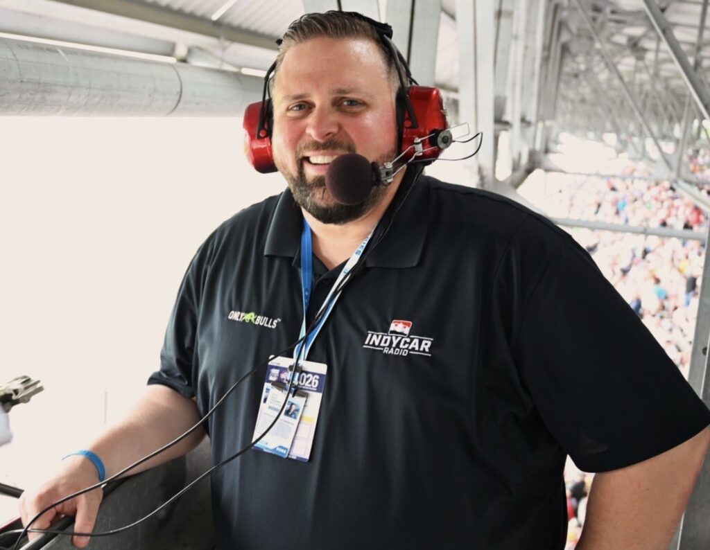 A radio broadcaster wearing a headset and microphone smiles from an elevated broadcast booth overlooking a racetrack filled with spectators. The person wears an INDYCAR Radio polo shirt and credential badge while preparing to cover the race.