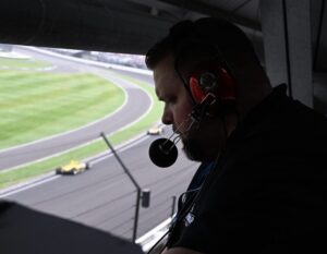 A radio broadcaster wearing a headset and microphone looks down at notes while working from a broadcast booth overlooking an active racetrack. Race cars are visible on the track in the background as the broadcaster focuses on the event coverage.