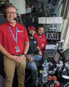 Three radio broadcasters wearing headsets work inside a motorsports broadcast booth overlooking a racetrack. One broadcaster stands smiling in the foreground while two colleagues sit at a desk with laptops, race timing monitors, notes, and broadcasting equipment as they prepare to cover the event.