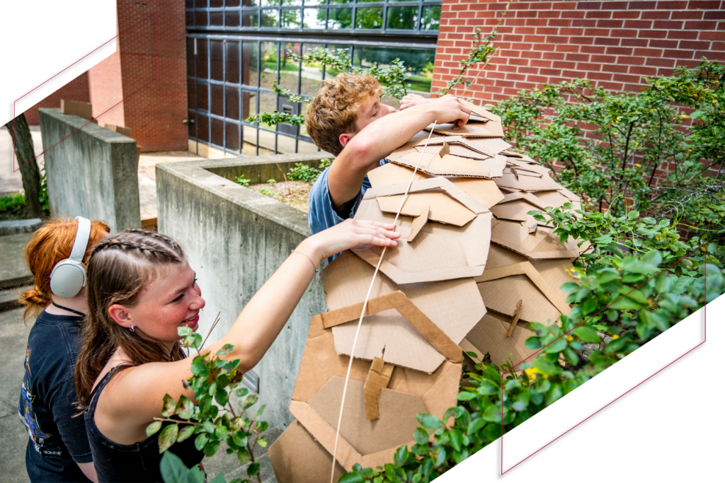 Two Ball State students work together outdoors to construct a large, layered cardboard structure among campus landscaping.