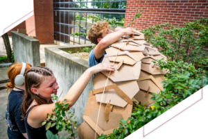 Two Ball State students work together outdoors to construct a large, layered cardboard structure among campus landscaping.