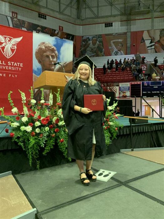 A person in a black graduation gown and cap holds a red folder on a stage with floral arrangements. A Ball State University banner is visible.