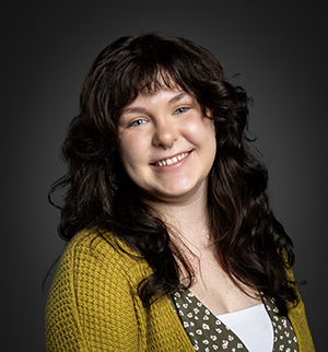 Headshot of a person with long, dark curly hair and a yellow cardigan, smiling at the camera against a dark gray background.
