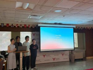 four students standing in front of a projector screen displaying the title slide of a Powerpoint presentation