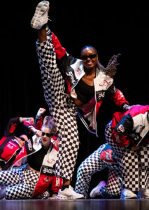 Woman is standing on stage holding one foot above her head doing the splits while standing up. There are several other women sitting around her in a pose. They are obviously all dancing and the photo caught them in motion, because you can see hair of the dancer that is standing flung out mid-motion. They are all wearing the same black and white checkered pants, white sneakers, and red, black, and white race car jackets with black tank tops under the jackets. 