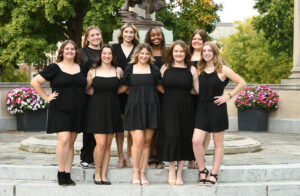 Nine college-age women stand in two rows posing and smiling to the camera. They are all in black dresses or black dress clothes. They each have their arms linked behind the person or people beside them. They are standing in front of the Beneficence statue on Ball State campus. 