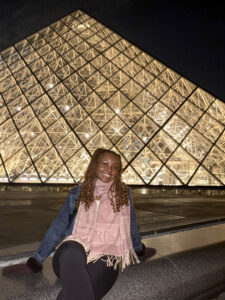 Top 100 Student Jamie Johnson-Harris sitting in front of the Louvre Museum in Paris, France. The building is a glass pyramid and lit up with lights. It is night time so the building is lit up bright behind her. 