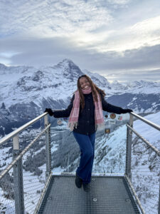 Top 100 Student Jamie Johnson-Harris is standing smiling and posed at a railing that overlooks what appears to be a range of mountains covered in snow. 