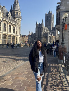 Jamie Johnson-Harris on a clear sunny day, though she is wearing a winter coat. She is smiling at the camera standing on a cobblestone street in front of what looks like a very old European town. 