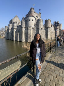Jamie Johnson-Harris standing in front of a railing on a cobble stone street. Behind her is what looks like a castle sitting in a river or moat. It is a sunny day, though Jamie has a coat on, and she is posed, smiling at the camera.