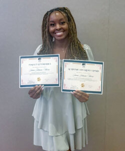 Top 100 Student Jamie Johnson-Harris standing posed agains a wall holding up two certificates, smiling at the camera. Both certificates were awarded in April of 2025 from the Ryan Family Scholars. One is for Perfect Attendance, and one is the Academic Excellence Award. 