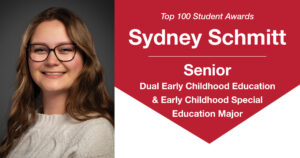 A woman with long, dark brown slightly curly hair hair wearing an off-white cable knit sweater and glasses smiles to the camera in front of a plain gray background. Text to the right reads, "Top 100 Student Awards Sydney Schmitt May 2026 Senior Dual Early Childhood Education & Early Childhood Special Education Major.”