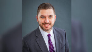 Head-and-shoulders portrait of a man smiling at the camera. He is wearing a gray suit jacket, white collared dress shirt, and purple plaid tie. He has brown hair, a mustache, and a beard with hints of gray, and is posed against a gray background.