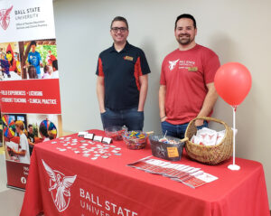 Two men stand behind a table covered with a red tablecloth featuring the Ball State University Teachers College logo. The table displays information packets, stacks of business cards, stickers, baskets of candy, and a red balloon attached to a stick. Beside the table is a large standing banner for the Ball State University Office of Teacher Education Services and Clinical Practice. The banner lists field experiences, licensing, student teaching, and clinical practice, and includes several photos of Ball State students, staff, and Charlie Cardinal interacting with children and students. Both men are wearing Ball State Teachers College shirts and jeans and are smiling at the camera.