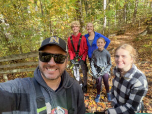 A father takes a selfie of his family while standing on a forest trail covered in orange fall leaves. He is wearing a gray sweatshirt, sunglasses, and a baseball cap with an “N” logo. Behind him stands the mother in a blue windbreaker alongside their three children. The oldest child, a boy with short curly blonde hair, wears a red sweatshirt and stands near his mother. In front of them is the youngest daughter in a gray jacket and purple pants. Closer to the camera is the middle child, a girl wearing a black-and-white plaid fleece jacket. The family is surrounded by trees with green and yellow leaves on a sunny autumn day, and everyone is smiling at the camera. The father and son also appear to be wearing backpacks or climbing gear.