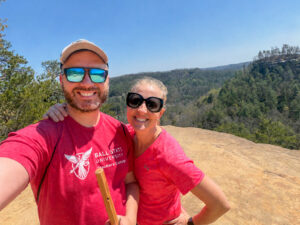 A man and woman take a selfie together while standing on a rock overlooking a tree-covered valley on a sunny day. Both are wearing sunglasses and red shirts, and the man’s shirt features the Ball State University Teachers College logo. He is also wearing a baseball cap and holding a hiking stick. The woman has her arm around the man’s shoulder, and both are smiling at the camera. Behind them is a bright blue sky with no clouds visible.