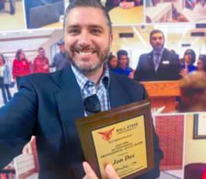 A man smiles at the camera while taking a selfie indoors. He is wearing a blue suit jacket over a blue-and-white plaid shirt that is unbuttoned at the top, revealing a white undershirt. A pair of sunglasses hangs from the open collar of his shirt. He has short brown hair and a trimmed mustache and beard with some gray mixed in. He is holding a plaque that reads, “Ball State University Teachers College 2025–2026 Outstanding Professional Staff Award Presented to Jon Dee in recognition of demonstrated achievement.” Behind him is a slightly blurred photo wall featuring an image of the same man speaking at a podium.