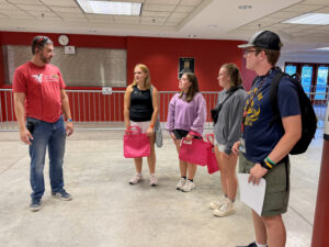 A man stands on the left side of the photo speaking with four Ball State students gathered beside him inside Worthen Arena. He is wearing jeans and a red T-shirt that reads “Ball State University Teachers College.” Sunglasses rest on top of his head, and a walkie-talkie clipped to his jeans is connected by a wire to an earbud in his ear. The group of students includes three young women and one young man. The women are each carrying red tote bags, while the young man has a backpack and is holding a piece of paper. They are standing on the lower level near the closed ticket office. Glass doors behind them lead outside, where sunlight can be seen coming through.