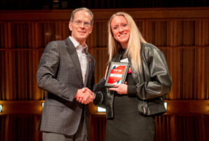 An older man and a young woman stand on stage shaking hands and smiling at the camera. The young woman is holding a plaque that reads “Top 100” and includes her name, Alex McPhee; the remaining text is too small to read. The man is wearing a gray plaid suit jacket with gray slacks and a white button-up dress shirt. The young woman is wearing a black dress with a black leather jacket and has long blonde hair.