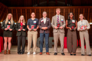Six young men and women stand on stage smiling and posing for the camera with the Director of Athletics at Ball State, who is standing in the center of the group. The student-athletes are each holding a plaque that reads “Top 100.”