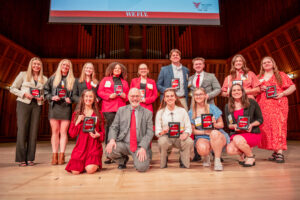 A group of 13 young men and women stand and kneel on stage, smiling and posing for the camera alongside an older man (the Dean of the College of Health) wearing a gray suit and red tie. The students are dressed in business casual attire and each is holding a plaque that reads “Top 100.”
