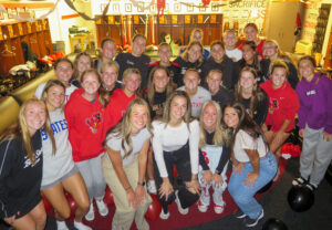 A large group of 27 young women stand together in a locker room, smiling and posing for the camera. The ceiling is decorated with streamers, and the group is dressed casually in athletic wear.