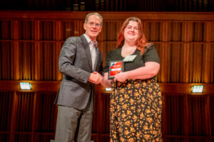 Elizabeth Frame stands on stage shaking hands with Ball State President Geoffrey Mearns after receiving her Top 100 Student Award plaque, which she is holding in her left hand. Both Elizabeth and President Mearns are smiling toward the camera. Elizabeth is wearing a dress with a black top and a black skirt featuring a yellow floral pattern. The photo captures the moment she is recognized for being named one of Ball State University's Top 100 Students.