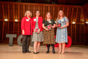 Four women stand on a stage, smiling and posing for a photo in front of large letters spelling “Top 100.” On the far left is Teachers College Associate Dean Susan Tancock, wearing a red jacket over a black shirt and black pants. Standing beside her are three Ball State students who were recognized as Top 100 Student Award recipients and attended the celebration ceremony honoring all award winners. Each student is holding a plaque recognizing the achievement. Elizabeth Frame is the second woman from the right and is wearing a dress with a black top and a black skirt featuring a yellow floral print. The group smiles toward the camera while celebrating their recognition as outstanding Ball State students.