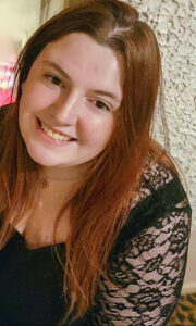 Close-up selfie of Elizabeth Frame smiling at the camera. She is wearing a black dress with lace sleeves, and her long red hair is worn down. The photo is taken against a plain white textured wall, creating a simple background that keeps the focus on her face.