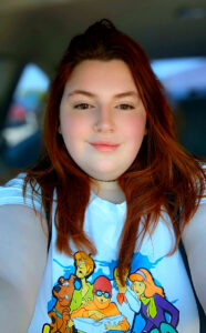 Selfie of a woman taken inside a car with a blurred background. She has long red hair and is wearing a white T-shirt featuring cartoon characters from Scooby-Doo.