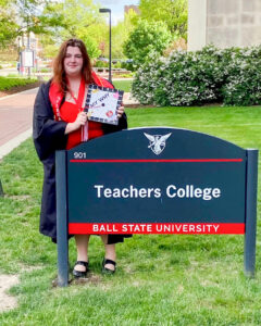 Elizabeth Frame stands outdoors on a sunny spring day beside the Teachers College sign on Ball State University’s campus. She is wearing a red dress, a black commencement gown, a red stole that reads “First Generation,” and a gold medallion on a red ribbon. She holds her decorated graduation cap in front of her. The cap features the alphabet around the border, the words “My Why” in the center, and signatures from students in the classroom where she completed her student teaching. Elizabeth has long red hair and is smiling beside the sign, which reads “Teachers College Ball State University” and displays the Beneficence logo.
