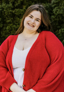 Portrait of Serenity Dutton, a young woman with long brown hair, standing outdoors in front of evergreen foliage. She is smiling at the camera with her head tilted to the side and is wearing a white shirt and a bright red cardigan. The image is cropped closely around her upper body, with the background softly visible behind her.