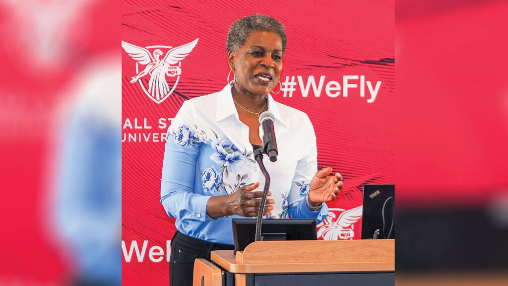 Renee Akins stands at a podium delivering remarks in front of a Ball State University backdrop featuring the University logo, the word “Welcome,” and the hashtag #WeFly over a pattern of cardinal red feathers. She has short dark hair with touches of white and wears hoop earrings and a necklace. Her collared button-up shirt transitions from white at the top to blue below, separated by a border of blue and white floral designs. She pairs the shirt with black pants as she addresses the audience.