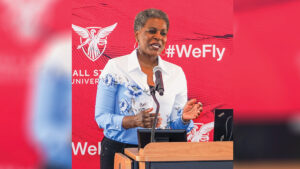 Renee Akins stands at a podium delivering remarks in front of a Ball State University backdrop featuring the University logo, the word “Welcome,” and the hashtag #WeFly over a pattern of cardinal red feathers. She has short dark hair with touches of white and wears hoop earrings and a necklace. Her collared button-up shirt transitions from white at the top to blue below, separated by a border of blue and white floral designs. She pairs the shirt with black pants as she addresses the audience.