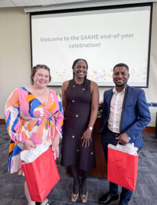 Three people stand smiling in front of a projection screen displaying the message, “Welcome to the SAAHE End-of-the-Year Celebration!” Renee Akins, scholarship donor and SAAHE alumna, stands in the center between the two recipients of the Renee Akins Scholarship. The students on either side of her hold large red gift bags with “Congratulations” handwritten on the front.The student on the left wears a vibrant long-sleeved wrap dress featuring pink, blue, yellow, white, and black patterns and has shoulder-length brown hair secured away from her face with small clips. Akins wears a predominantly black sleeveless dress with a subtle floral pattern and has long black braids pulled back from her face. The student on the right wears dark blue dress pants, a blue patterned sport coat with black trim, and a white dress shirt. He has short black hair and a neatly trimmed beard and mustache. SAAHE, which stands for Student Affairs Administration in Higher Education, is a master's degree program at Ball State University.