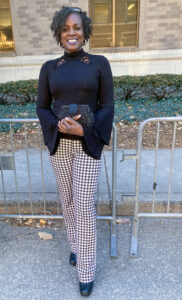 Renee Akins stands outdoors, smiling and posing for the camera in front of a brick building with tan and brown tones and a white stone base. The building features long horizontal windows and a row of low hedges. A sidewalk runs behind her, with a curb and metal fence positioned just behind her. Akins has short, curly black hair pulled back with sunglasses resting on top of her head. She wears gold hoop earrings and a high-neck top with long trumpet sleeves and small decorative cutouts near the neckline. She pairs the top with seersucker-patterned pants and black shoes and holds a black feathered and beaded clutch purse.