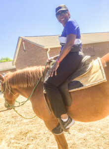 Renee Akins sits on a light brown horse in a saddle, smiling toward the camera on a bright, sunny day. She wears a black baseball cap, sunglasses, a purple shirt, black pants, and black sneakers, along with a watch featuring a red band. She appears to be holding an item tucked under her arm that may be a bag. In the background are two red brick buildings, and the ground is soft, well-trodden dirt, suggesting an outdoor riding or stable area.