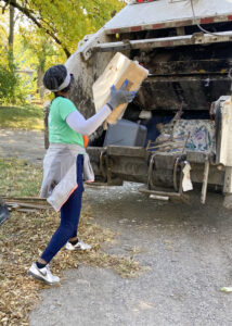 Renee Akins is pictured from behind as she prepares to toss a cardboard box into a garbage truck during a Keep Indy Beautiful cleanup event. The truck already contains assorted debris, including a large plastic container and an item with a white base and purple and green floral pattern. Additional litter is visible on the ground nearby.In the background, dense green trees partially obscure a bridge structure. Akins wears her dark hair pulled back with a gray headband, a gray long-sleeve shirt under a green T-shirt, gray work gloves, jeans, and white sneakers. A gray zip-up sweatshirt is tied around her waist, suggesting she had removed it earlier due to the heat.