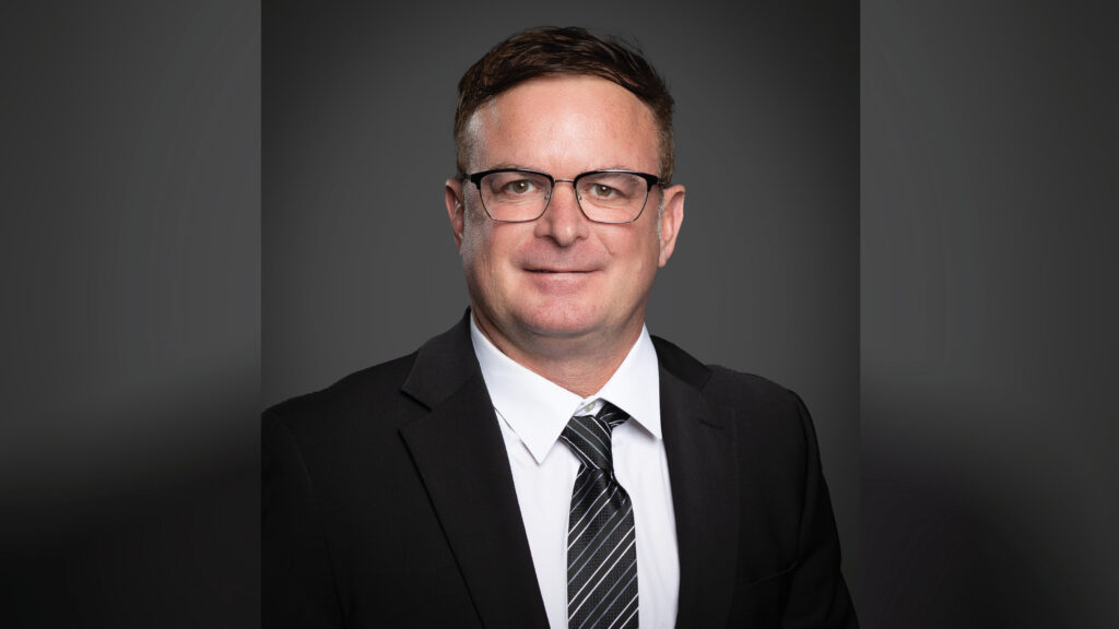 Head-and-shoulders portrait of David Roof wearing a black suit, white dress shirt, and black tie with gray and white stripes. He has short hair and black-rimmed glasses and is photographed against a dark gray studio backdrop.