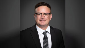 Head-and-shoulders portrait of David Roof wearing a black suit, white dress shirt, and black tie with gray and white stripes. He has short hair and black-rimmed glasses and is photographed against a dark gray studio backdrop.