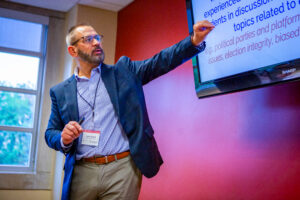 John Snoad of the Sphere Education Initiative stands at the front of a room during the 2024 Civic Learning Symposium, pointing toward a monitor displaying text while speaking. He wears a navy blazer, blue patterned dress shirt, and tan pants, and holds a pen with a lanyard and name badge around his neck. He has short brown hair, a mustache and beard with some gray, and black-framed glasses. The room has a red wall behind the monitor, a tan adjacent wall, and a window showing green trees.