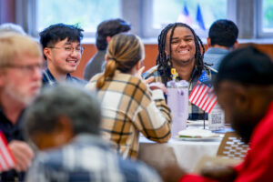 Three young adults sit at a round table in the L.A. Pittenger Student Center Ballroom during the Civic Learning Symposium, smiling and talking. A water bottle and a small American flag are on their table. Other attendees seated at nearby tables are visible in the foreground and background, slightly out of focus, along with windows and additional American and Indiana flags.