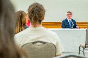 David Roof sits at a panel table at the front of a room during a discussion, speaking to an audience. The photo is taken from the audience perspective, with the backs of attendees’ heads visible in the foreground. He wears a blue suit, white dress shirt, and striped, blue tie. A microphone on a short stand sits on the white tablecloth-covered table in front of him. The background features wood paneling on the lower wall and a plain white upper wall.