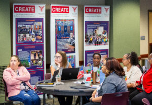 Six women sit at a round table in a room with green and tan walls during a CREATE event at Ball State University’s Teachers College. Behind them are three CREATE banners reading “Civic Renewal through Education for Agency, Tolerance, and Engagement,” with additional headings including Civic Engagement, Civic Agency, and Civic Knowledge. Several participants have laptops, notebooks, and drinks on the table, along with a small U.S. flag. Some women appear to be listening to a speaker off-camera while others are in discussion.