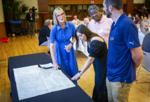 Four people gather around a table displaying a copy of the United States Constitution during the Civic Learning Symposium in the L.A. Pittenger Student Center Ballroom. One woman leans over to examine the document while another gestures toward it as they talk. In the background, attendees are seated at round tables throughout the ballroom, and a videographer records the event from the back of the room.