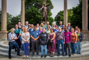 Twenty-seven participants in a STEM Teacher Workshop pose on the steps in front of the Beneficence statue on Ball State University’s campus. The statue and surrounding pillars are visible behind them, with green, leafy trees in the background. Each participant wears a name tag, though the details are not legible.