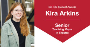 Header graphic for a Top 100 Student Awards blog post. On the left is a photo of Kira Arkins, a young woman with long red hair, standing indoors beside a metal railing with one hand resting on it. She is smiling at the camera and wearing a black T-shirt layered under a light green collared button-up shirt. The background is softly blurred. On the right side of the graphic, text reads: “Top 100 Student Awards,” “Kira Arkins,” “Senior,” and “Teaching Major in Theatre.”