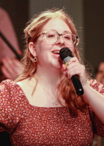 Close-up photo of Kira Arkins singing on stage while holding a microphone close to her mouth. She is wearing glasses, and her long red hair is partially pulled back from her face with the rest falling over her shoulders. She is dressed in a muted red square-neck top with a small white floral pattern. The background is softly blurred, keeping the focus on her as she performs.