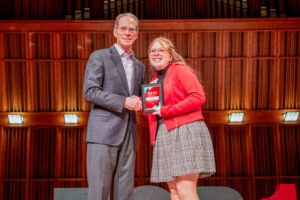 Kira Arkins stands on stage shaking hands with Ball State President Geoffrey Mearns after receiving her Top 100 Student Award plaque, which she is holding in her left hand. Both Kira and President Mearns are smiling toward the camera. Kira is wearing an above-the-knee black-and-white plaid skirt, a black turtleneck, and a red zip-up jacket. The photo captures the moment she is recognized as one of Ball State University's Top 100 Students.