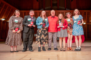 Seven people stand on stage during the Ball State Top 100 Student recognition ceremony. In the center is former College of Fine Arts Dean Seth Beckman, surrounded by the six College of Fine Arts students who were recognized as Top 100 Students and attended the ceremony. All are dressed in business casual attire. Kira Arkins is standing immediately to Dean Beckman's left (third from the right), wearing an above-the-knee black-and-white plaid skirt, a black turtleneck, and a red zip-up jacket. She is smiling and holding her Top 100 Student Award plaque as the group poses for the photo.